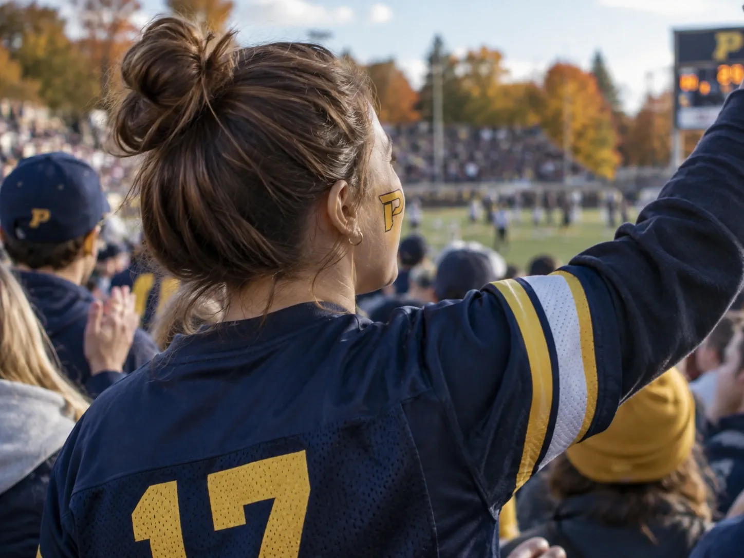 A fan in a navy number 17 jersey with a gold P painted on her cheek raises a fist in the stands, the field and an autumn tree line beyond.