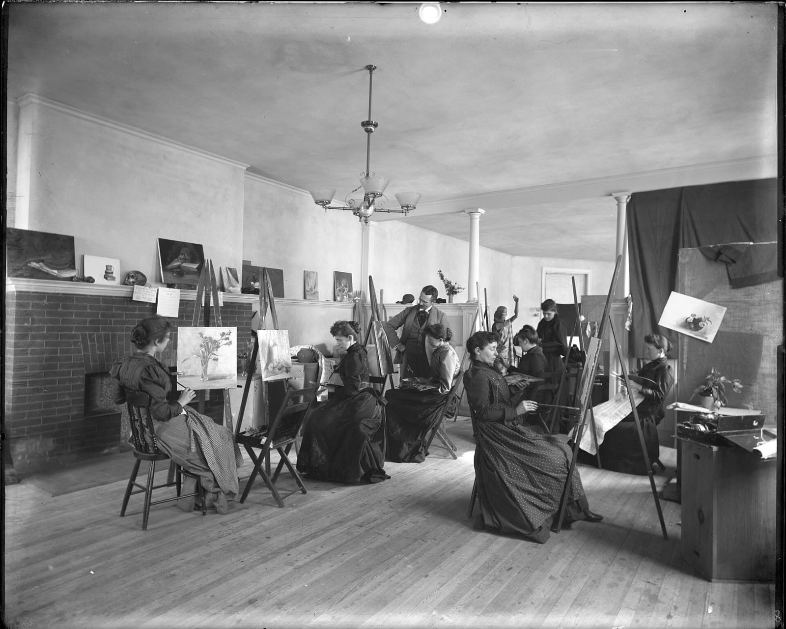 A black-and-white photograph of a late-nineteenth-century painting class: women in long dark dresses seated at easels painting still lifes while an instructor in a suit looks on.