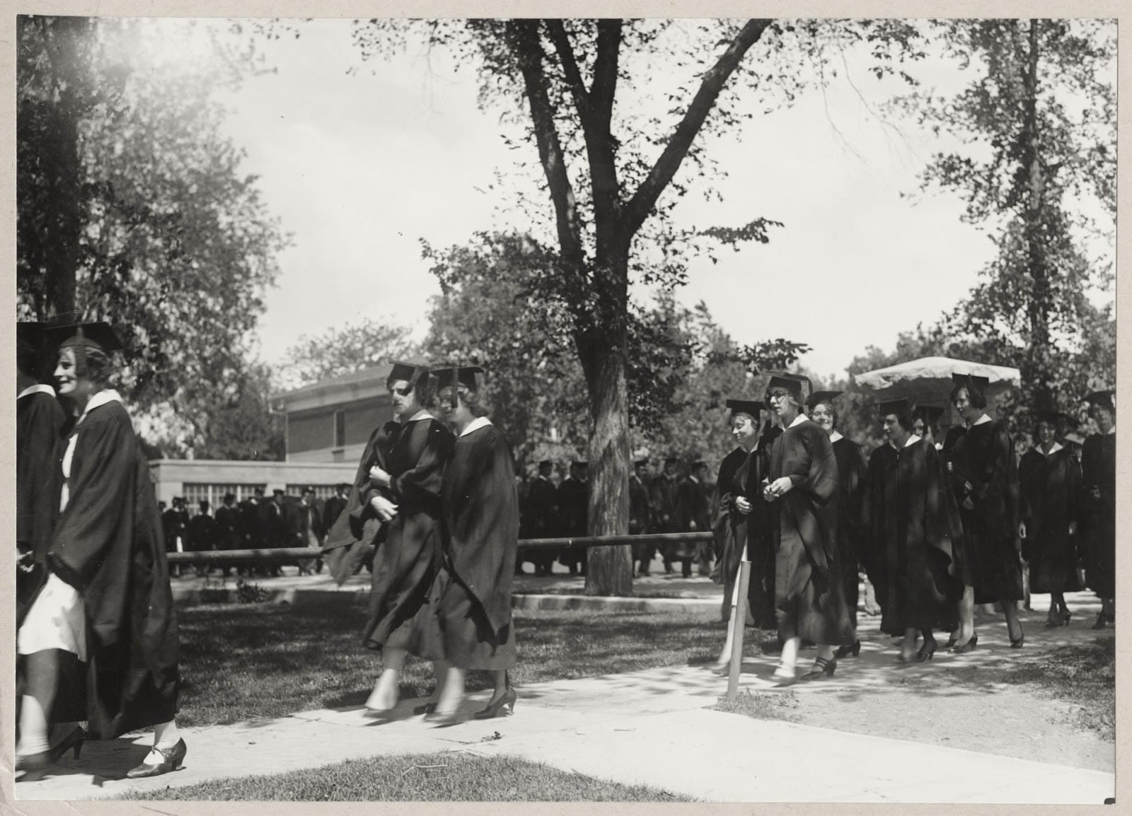 A historic photo of graduates in caps and gowns processing across campus.