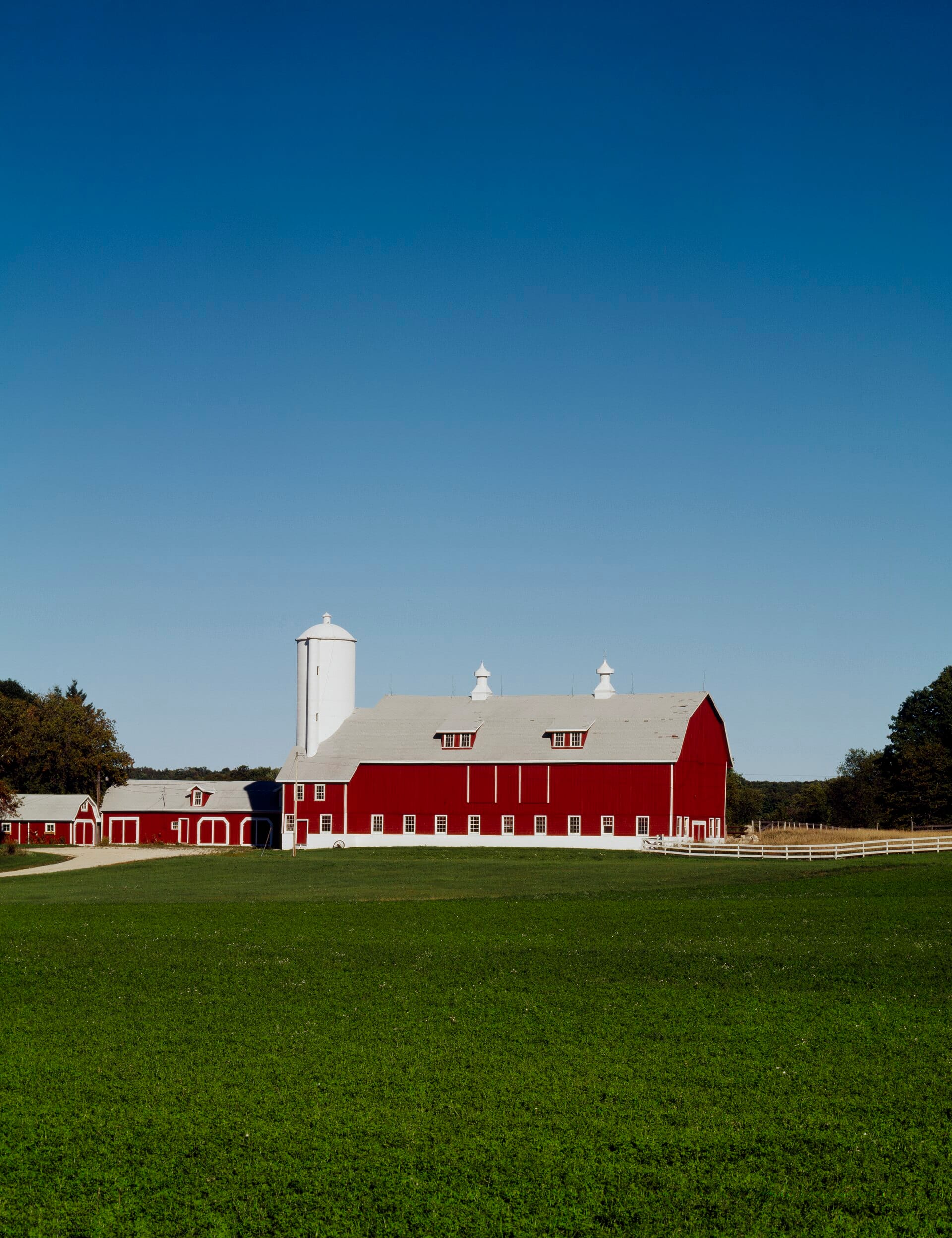 A long red barn with a white silo beside a green field under a clear blue sky.