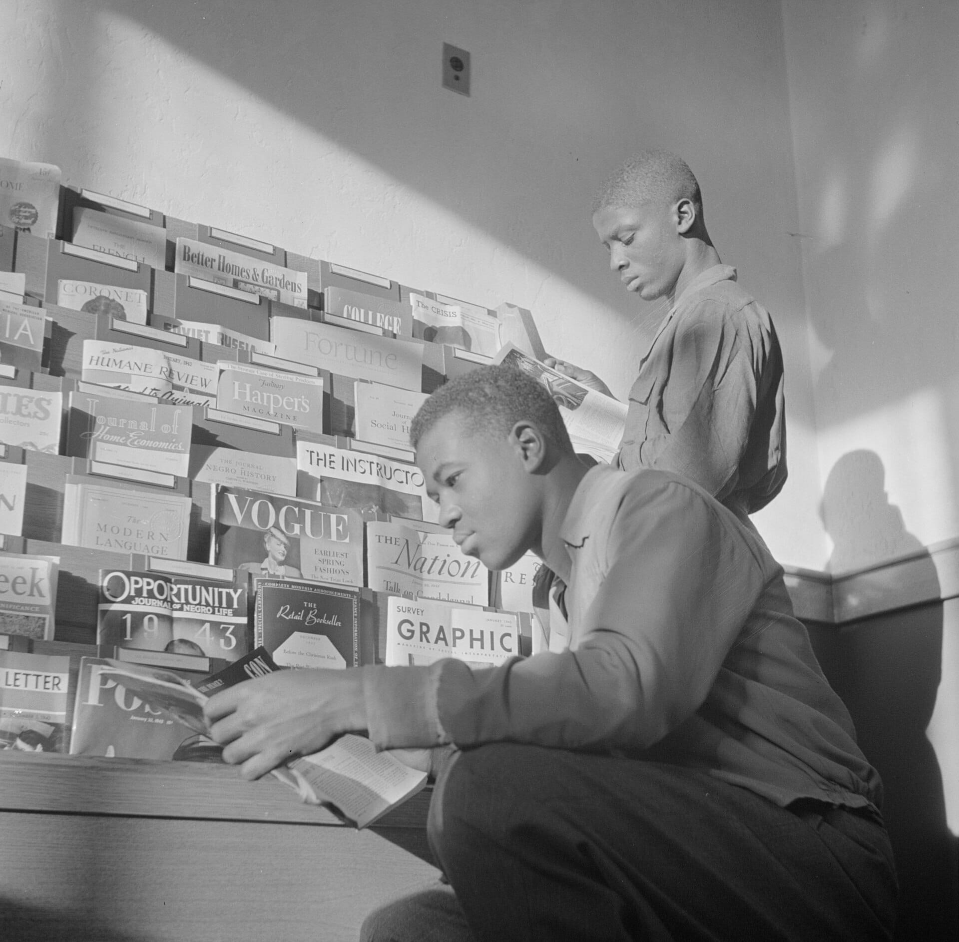 A historic black-and-white photo of two young men reading magazines from a library periodicals rack.