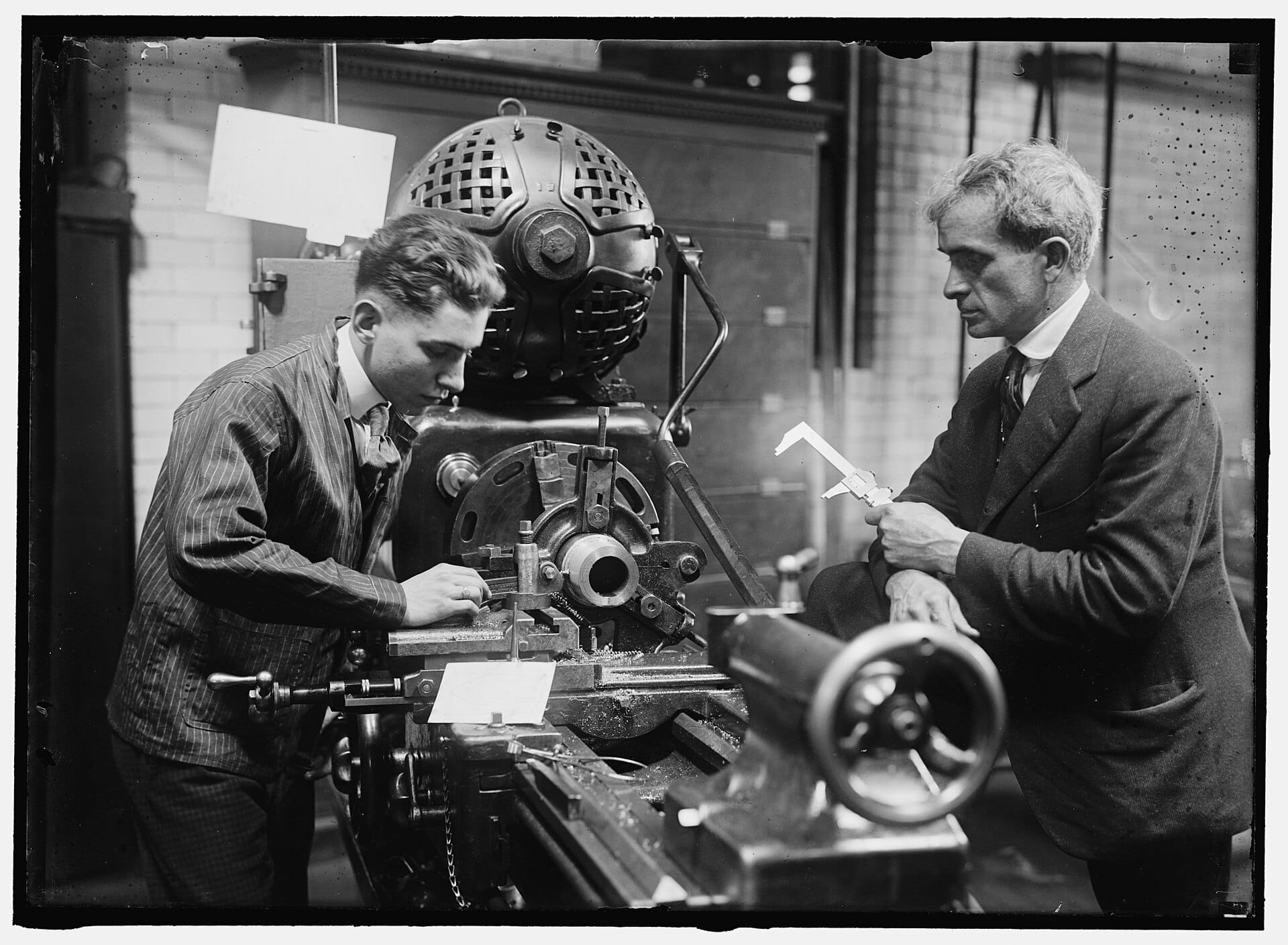A historic black-and-white photo of a young man working at a lathe while an instructor in a suit checks a part with calipers.