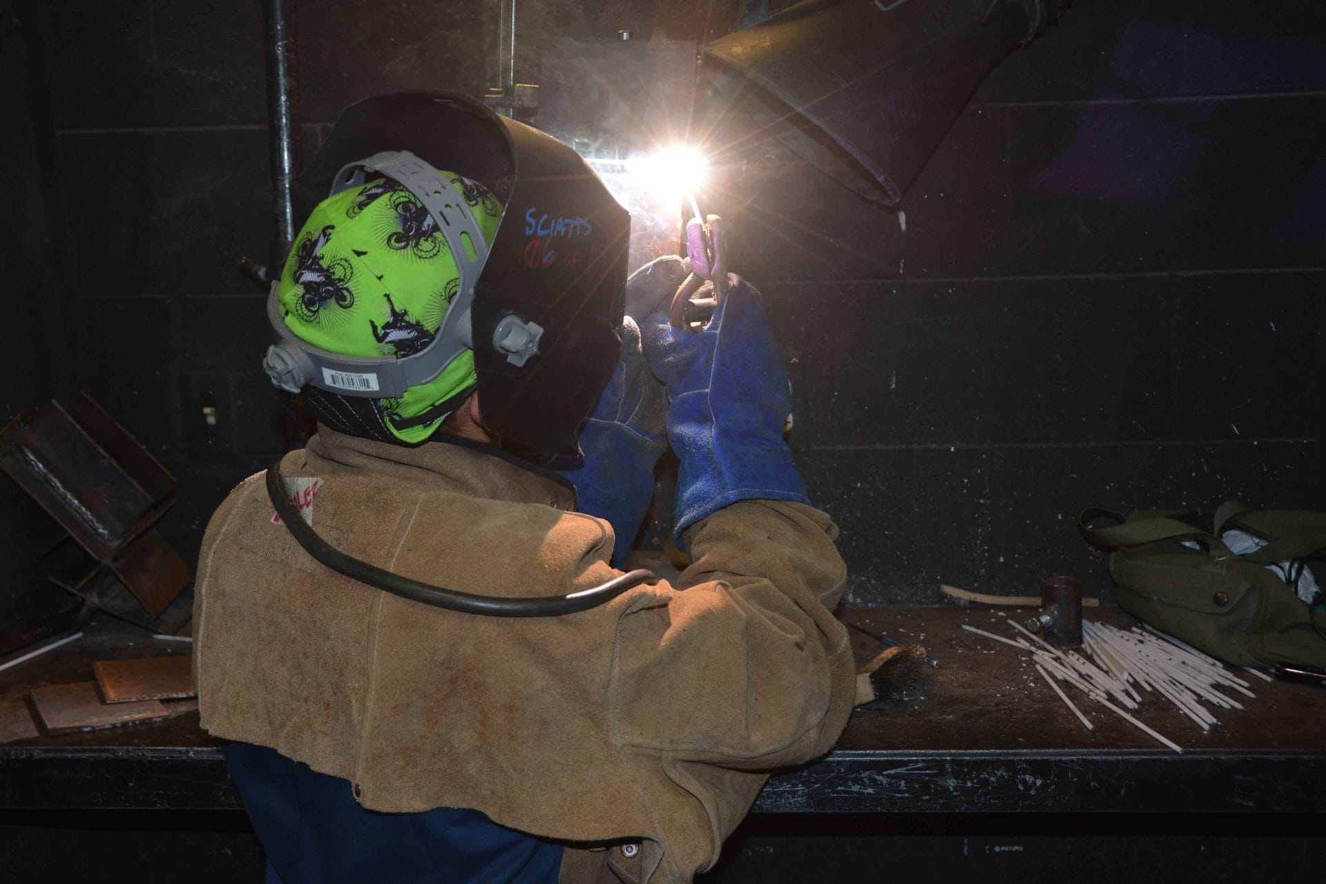 packaging technology program — A student welding at a bench, sparks lit against the dark.